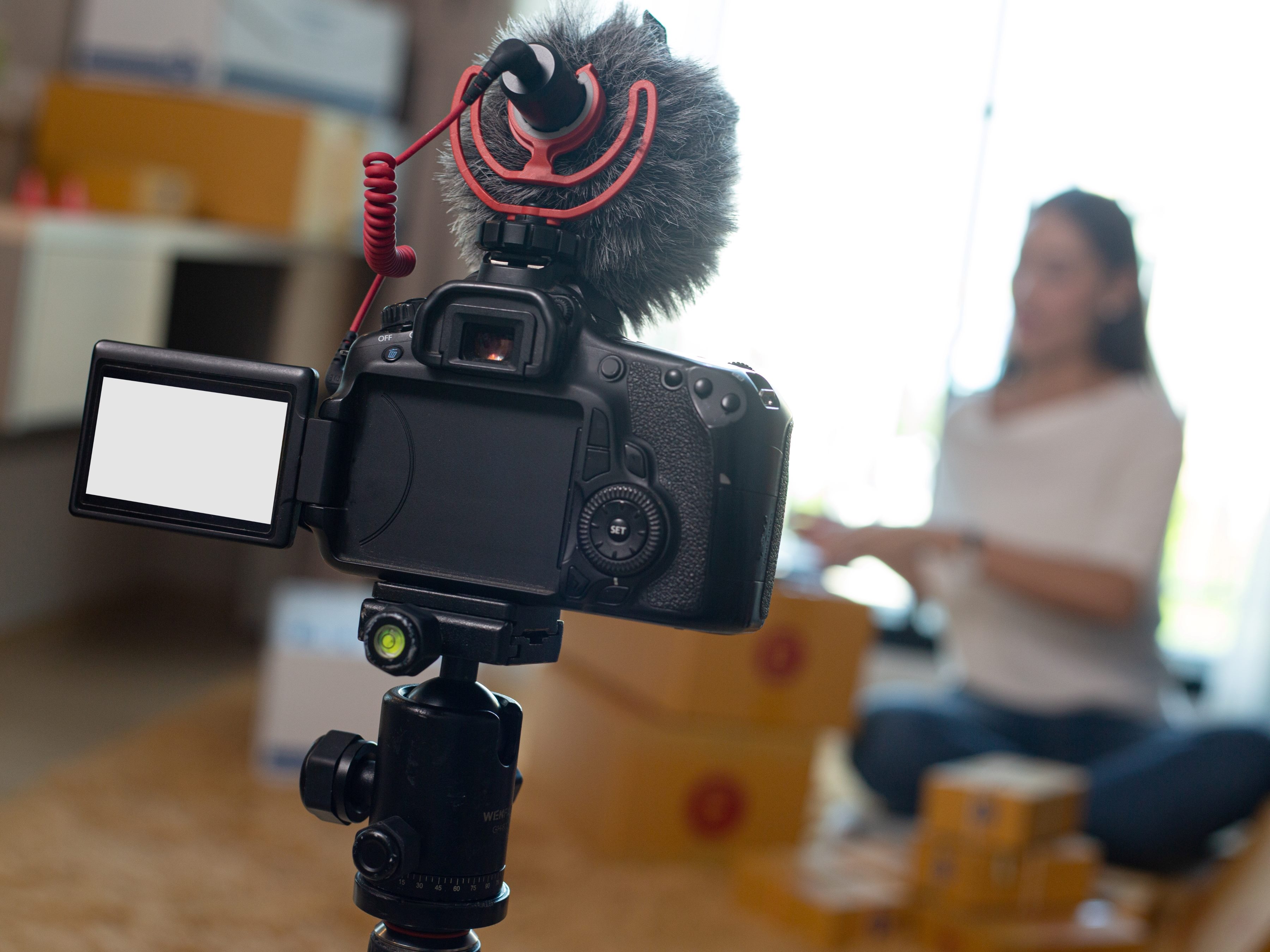Vlog recording for live video interview, with camera set up in foreground and person in background sitting on floor surrounded by boxes. 