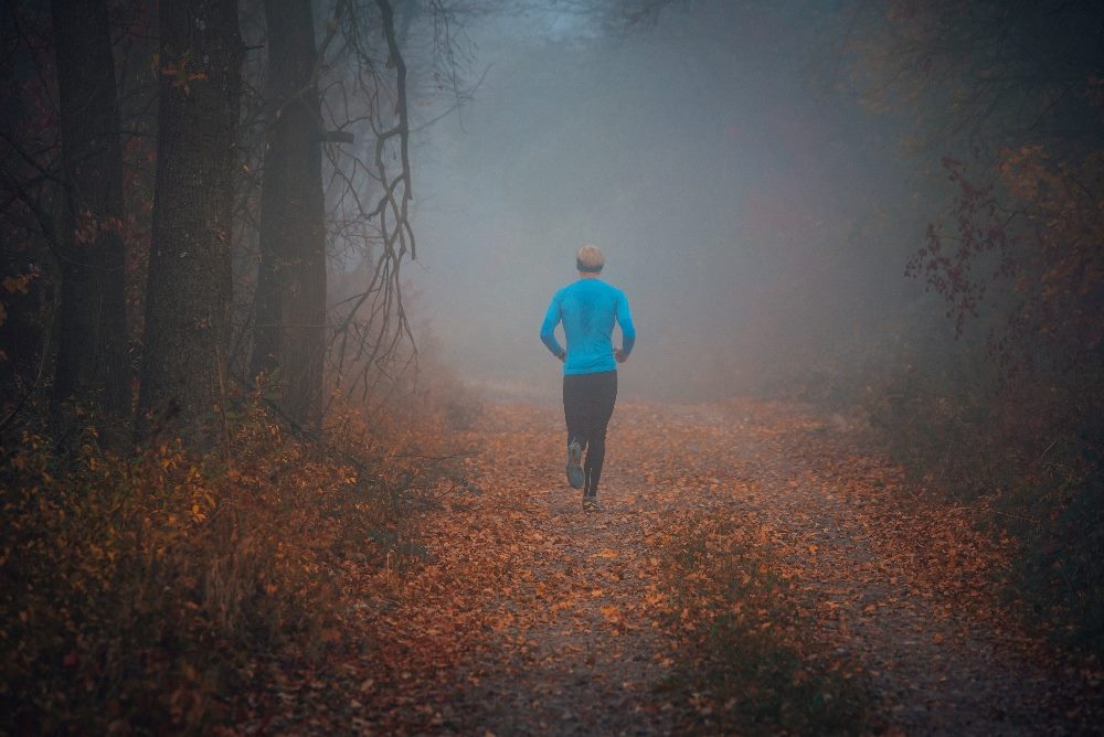 A lone runner, clad in a bright blue jacket, jogs away on a leaf-covered trail shrouded in a dense, foggy autumn forest. The image, symbolizing perseverance and exploration, subtly reflects the challenges and journey of generative AI implementation in uncharted territories.