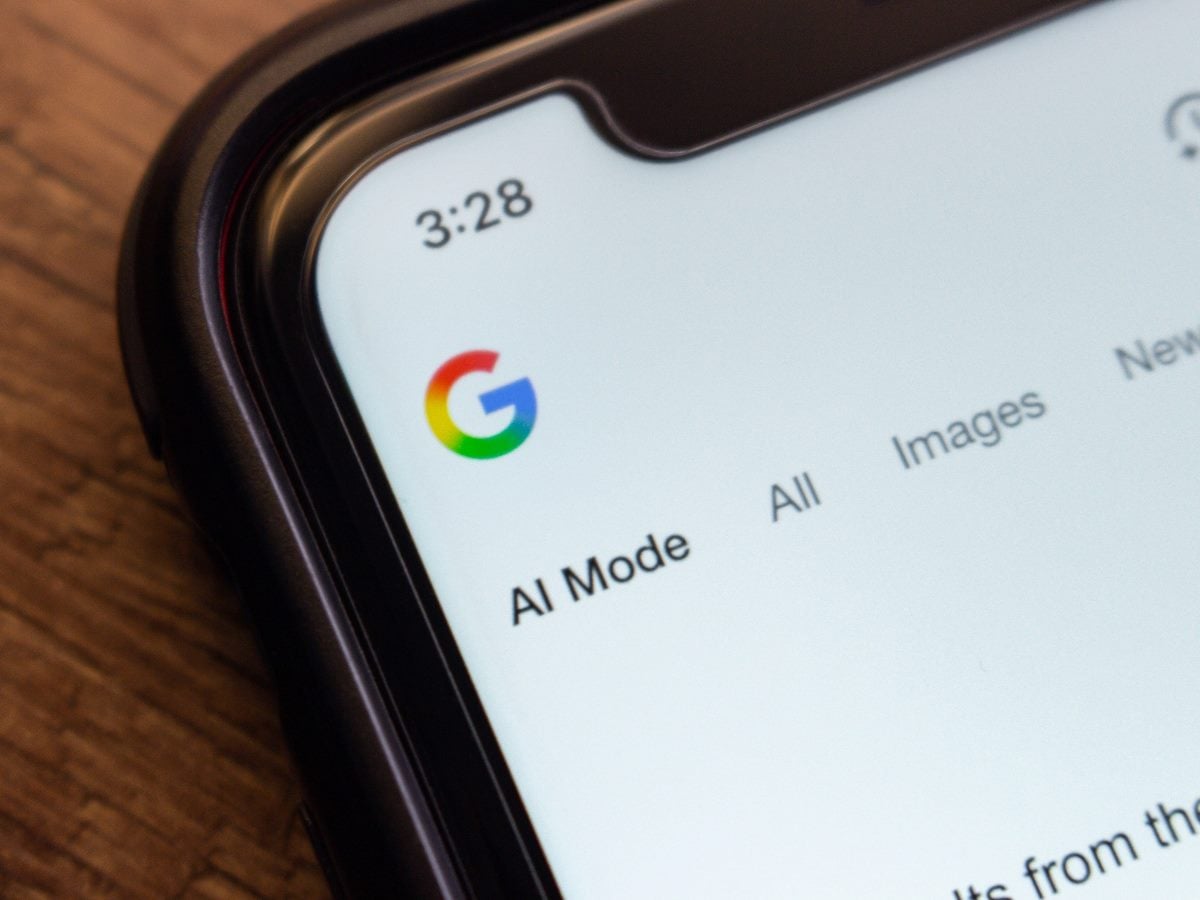 Close-up photo of a smartphone displaying the Google search interface with an “AI Mode” tab visible near the top navigation. The Google logo appears on the upper left of the screen, and the phone rests on a wooden surface.