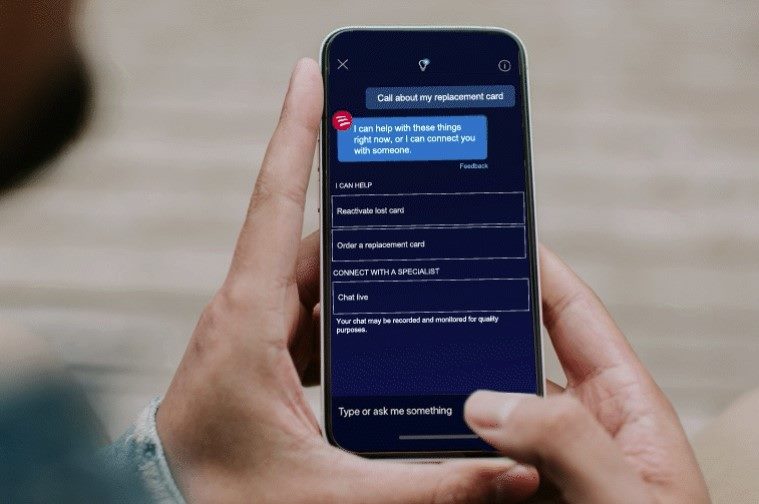 A person holds a smartphone showing the interface of Bank of America's AI-powered virtual assistant, Erica. The screen displays a chat with Erica, offering options such as reactivating a lost card, ordering a replacement card, or connecting with a specialist via live chat. The text input field at the bottom allows the user to type questions or commands.