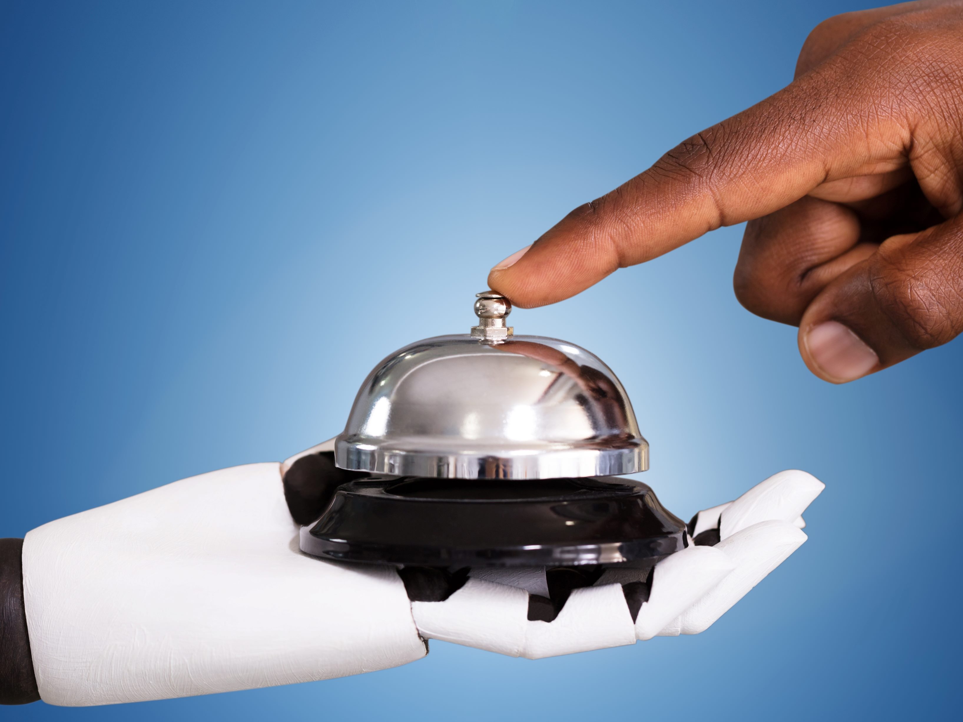 A white robot hand holds a service bell while a human hand points finger to ring it, suggesting the importance of AI in customer experience. 