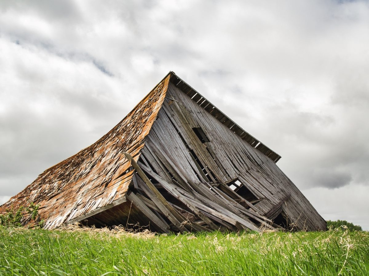 A weathered wooden barn with its roof and walls partially collapsed leans dramatically toward the ground in a grassy field. The aging structure shows broken boards, missing siding and open window gaps beneath a sky filled with thick gray clouds, illustrating severe structural deterioration and neglect.