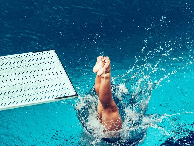 A female diver has taken the plunge from light blue diving board into the aqua-colored water of a swimming pool with pointed toes on entry suggesting the need for companies to take the risk of investing in AI in business.