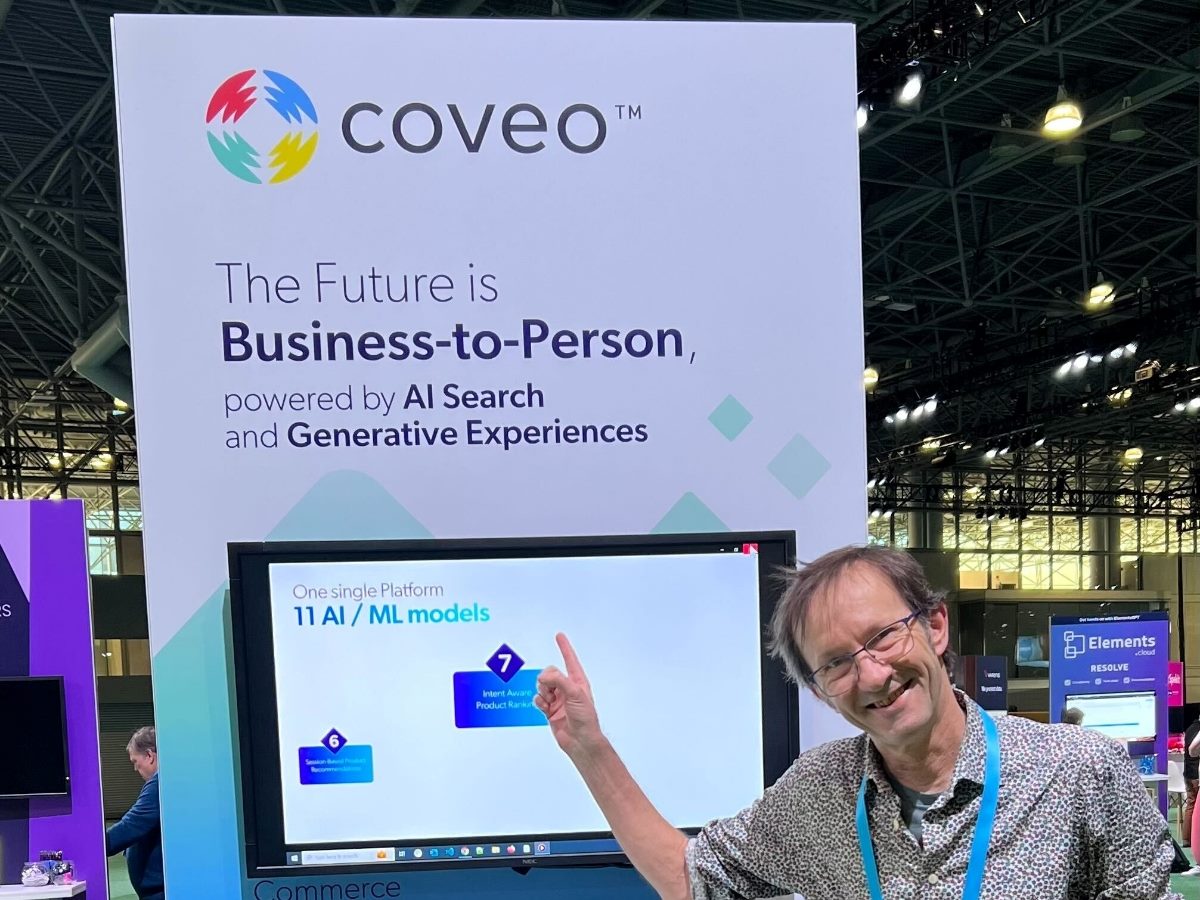 A man smiles and points toward a large Coveo booth display at an event. The sign behind him reads “The Future is Business-to-Person, powered by AI Search and Generative Experiences,” with screens and exhibitor booths visible in the background.