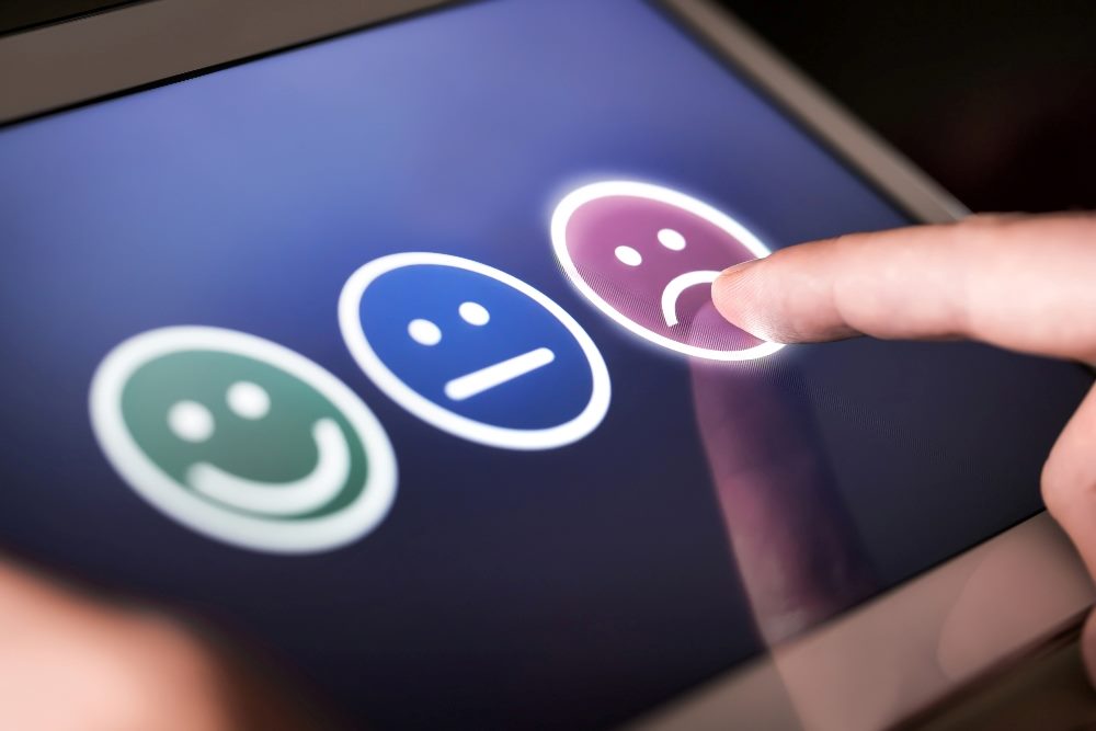 A man selects an unhappy face in online survey on a tablet indicating his confusion on disappointment in customers service, choices are a green happy face, a blue straight mouthed face and a purple unhappy face, suggesting challenges companies face in adopting AI customers service tools.