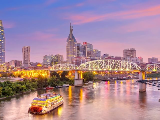 The Nashville, Tennessee, skyline flooded in pink twilight light with a river boat, a three-part bridge, tall downtown buildings, a river, and bright lights. 
