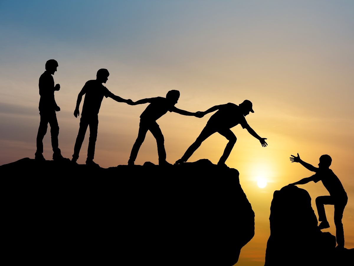 Silhouettes of several people standing on a cliff at sunset, reaching down to help pull another person up from a lower ledge, symbolizing collaboration, trust and support.