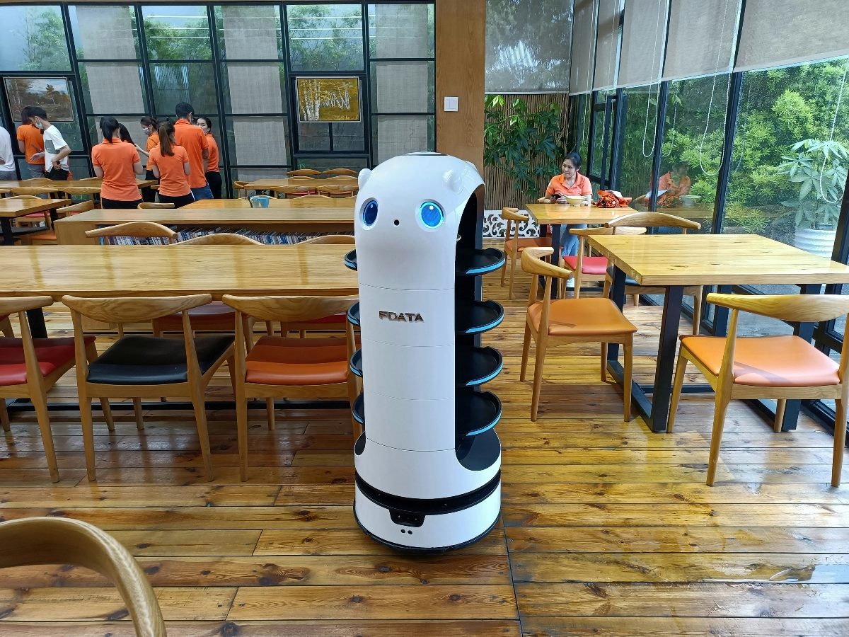 A robot kiosk machine in a work setting with people in the background wearing matching orange shirts.