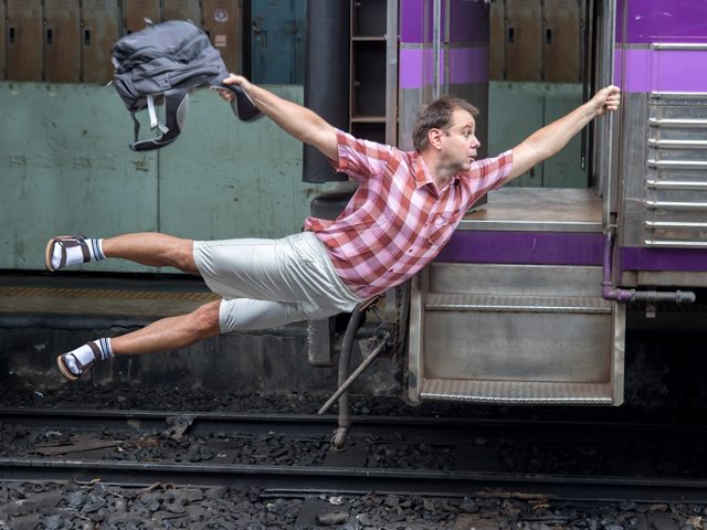 A light-skinned man wearing shorts and holding a backpack "flies" from the side of a train as if it is moving away at speed.