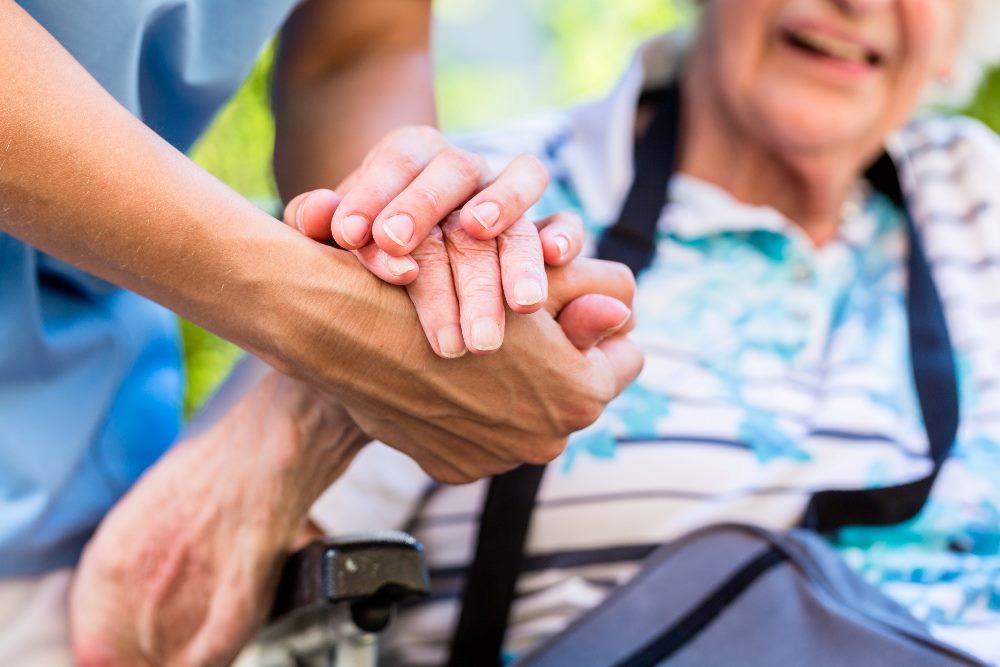 A nurse wearing scrubs holds the hand of and consoling a senior patient who is sitting in a wheelchair in piece about AI in customer experience in healthcare.