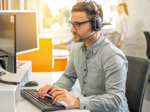 Male professional call center telesales agent wearing headset using computer in customer care support service office.