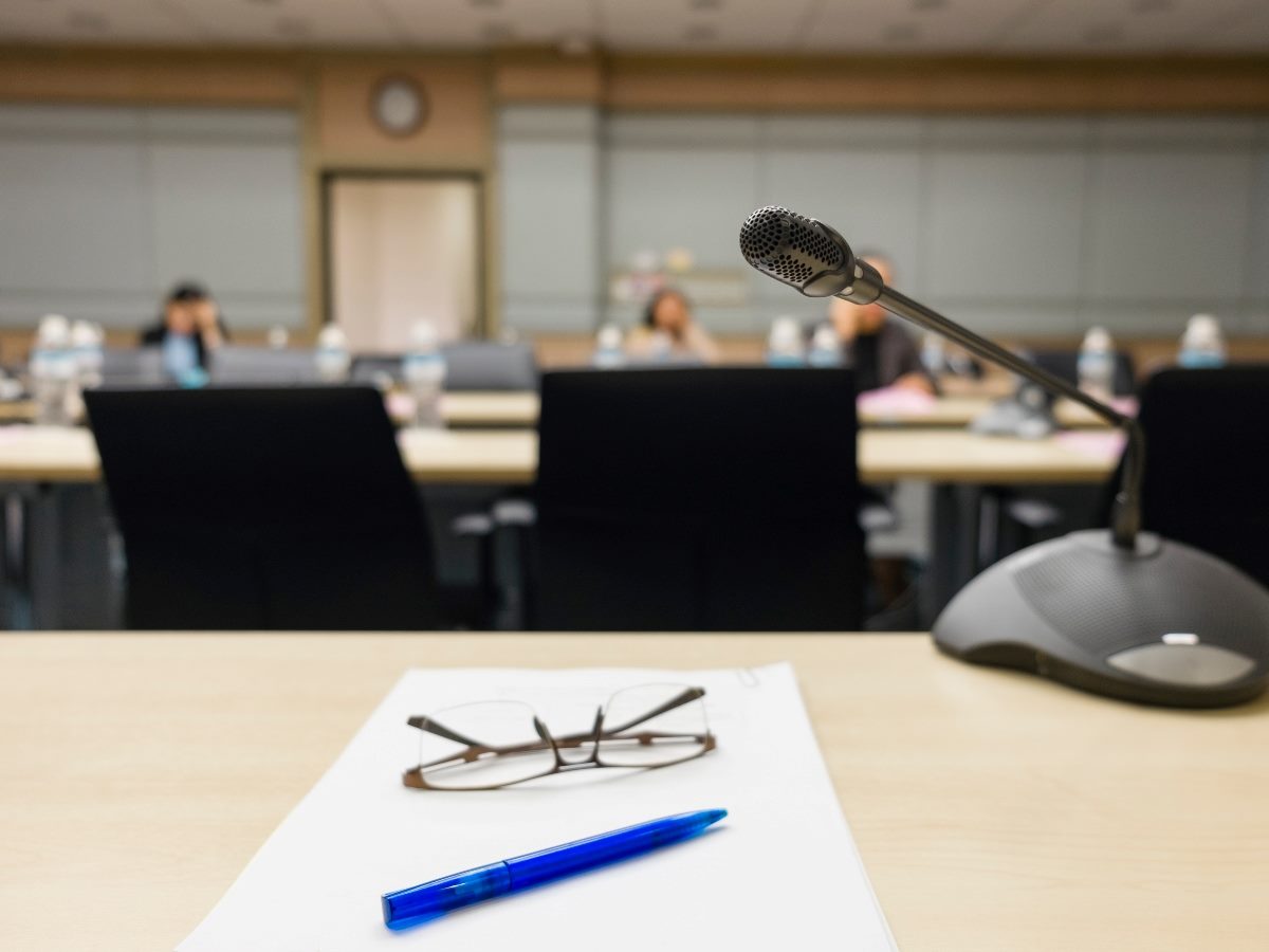 Close-up of a conference table with a microphone, eyeglasses, pen, and paper in focus, with blurred boardroom participants in the background.