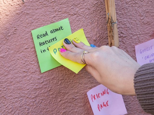 close up of a hand with brightly painted finger nails placing a sticky note in a rudimentary workflow