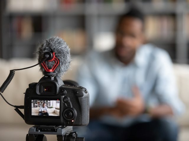 Video camera in foreground set up to film man sitting down in background.