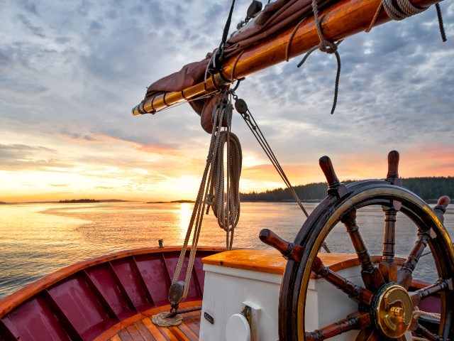  Close up of a schooner's wooden steering wheel, bow and boom against a dramatic sky at dawn.