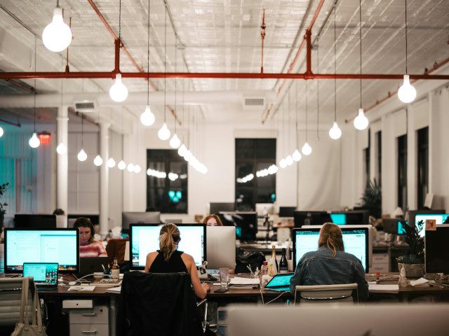 Employees work on computers at desks in an open office with hanging lights.