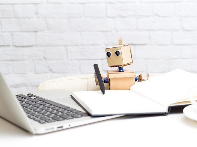 A small cardboard robot model with googly eyes holds a pen as if writing into a notebook, while also sitting at a Macbook on a desk.