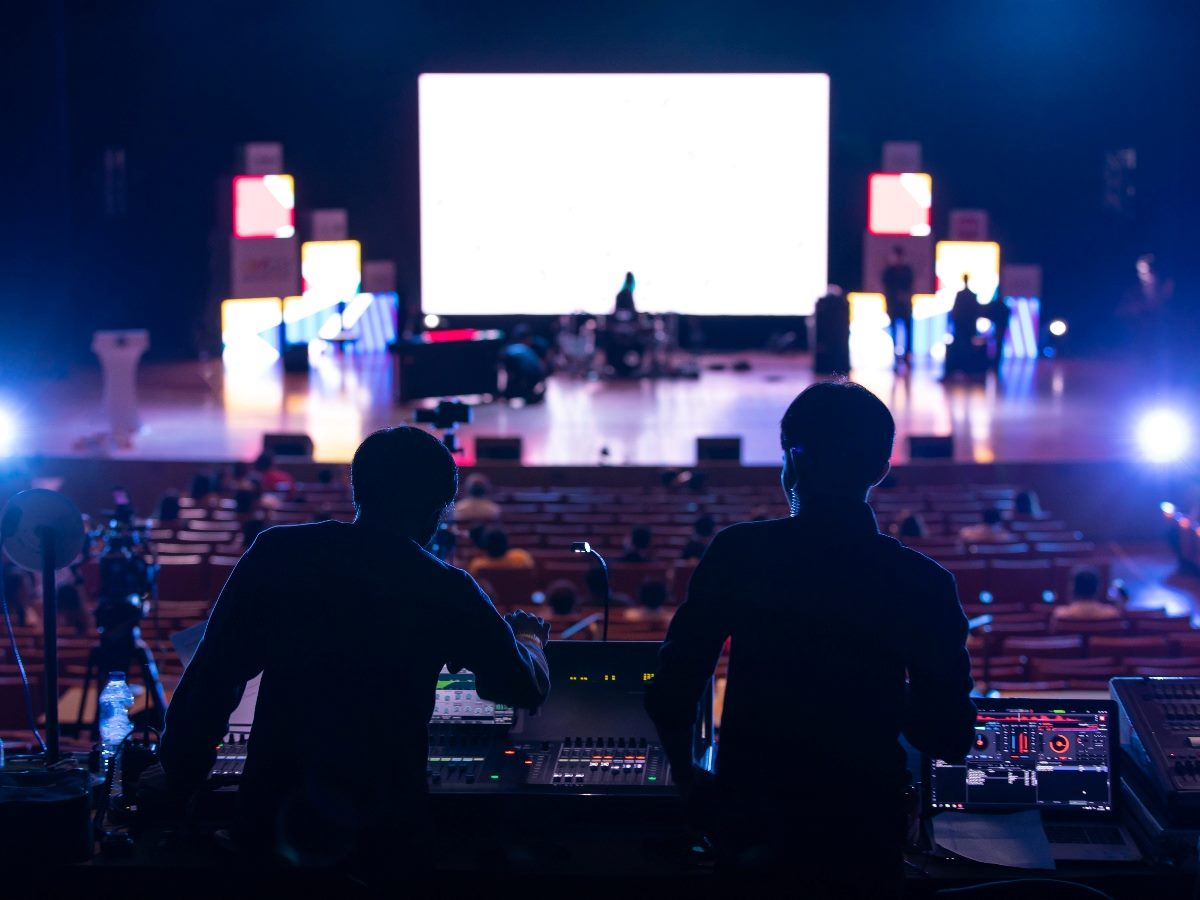Two audio-visual technicians operate sound and lighting equipment at the back of a dimly lit auditorium during a live event, with a brightly lit stage and digital screens in the background.
