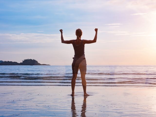 Woman with stretched up arms standing looking out toward the ocean at idyllic beach during sunset.