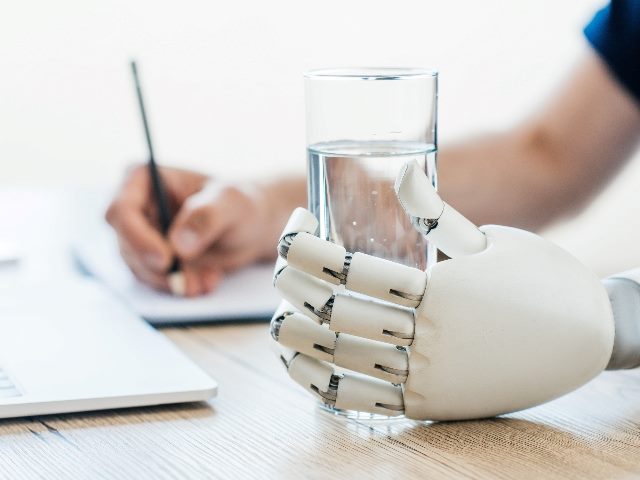 Selective focus on robotic arm holding glass of water while a human hand blurred in background takes notes at wooden table.