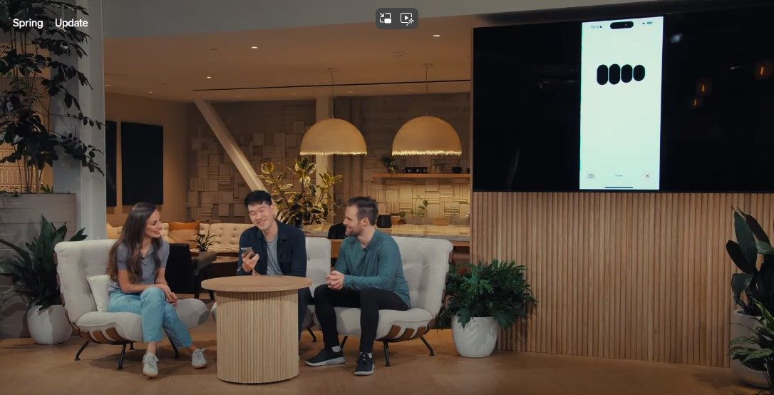 Three people are seated around a small table in a modern, well-lit room during OpenAI's Spring Announcement. They appear engaged in discussion, with one person holding a phone. Behind them, a large screen displays an image from the phone. The setting includes stylish furniture, plants, and warm ambient lighting, suggesting a relaxed yet professional atmosphere in piece about AI in customer service.