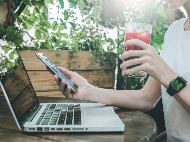 A woman sits outside at a table with trees in the background while working on her laptop while simultaneously checking her phone, suggesting the importance of seamless omnichannel strategies.
