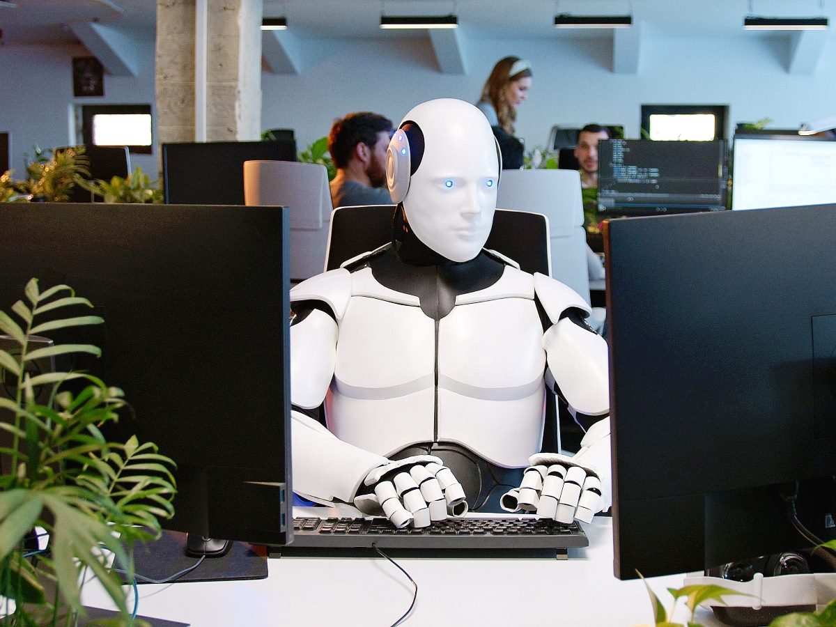 White robot sitting at table in office and typing on keyboard. 