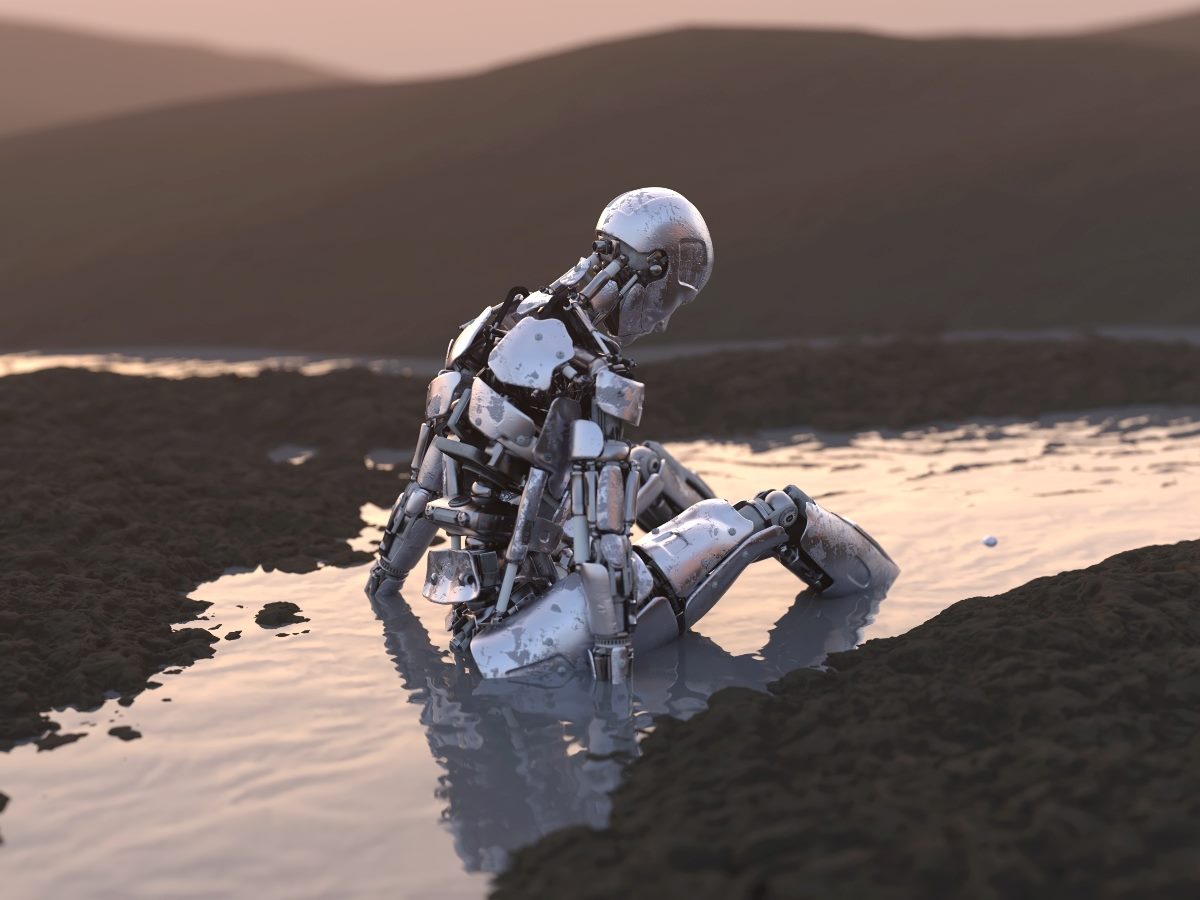 A humanoid robot sits partially submerged in shallow water, appearing stuck and motionless, with a barren landscape in the background at sunset.