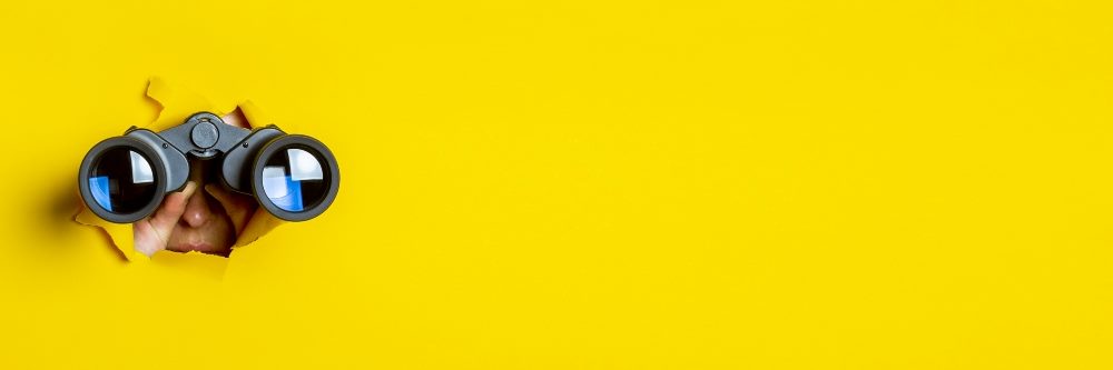 Female hand holds black binoculars on a yellow background.