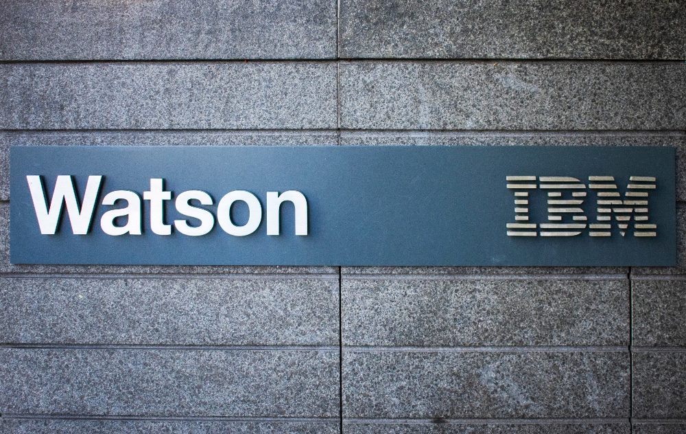 The names of IBM and Watson are in white lettering on a slate blue board on the gray stone facade of the IBM headquarters building in story about generative AI history.