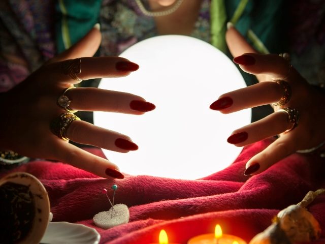 Hands of a female fortune teller around a crystal ball sitting on a red table cloth with candles in foreground