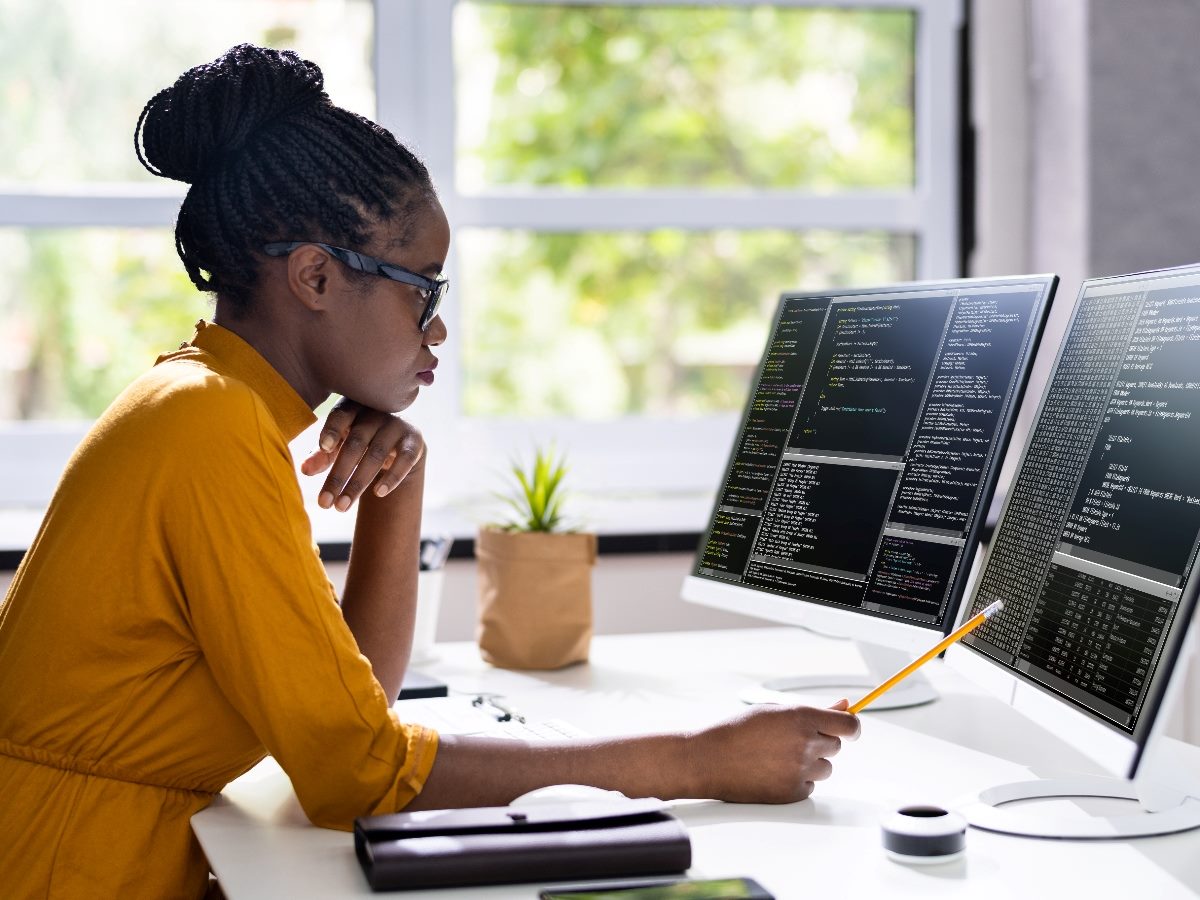 A woman sits at multiple computer terminals looking at screens filled with code.