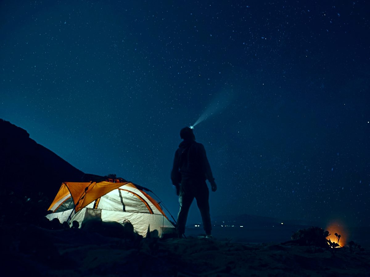 Man standing outside a tent at night with a headlight on looking into the night sky.