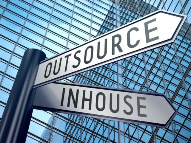 Street signs pointing different directions, one says outsource and the other says in-house, with building windows in the background.