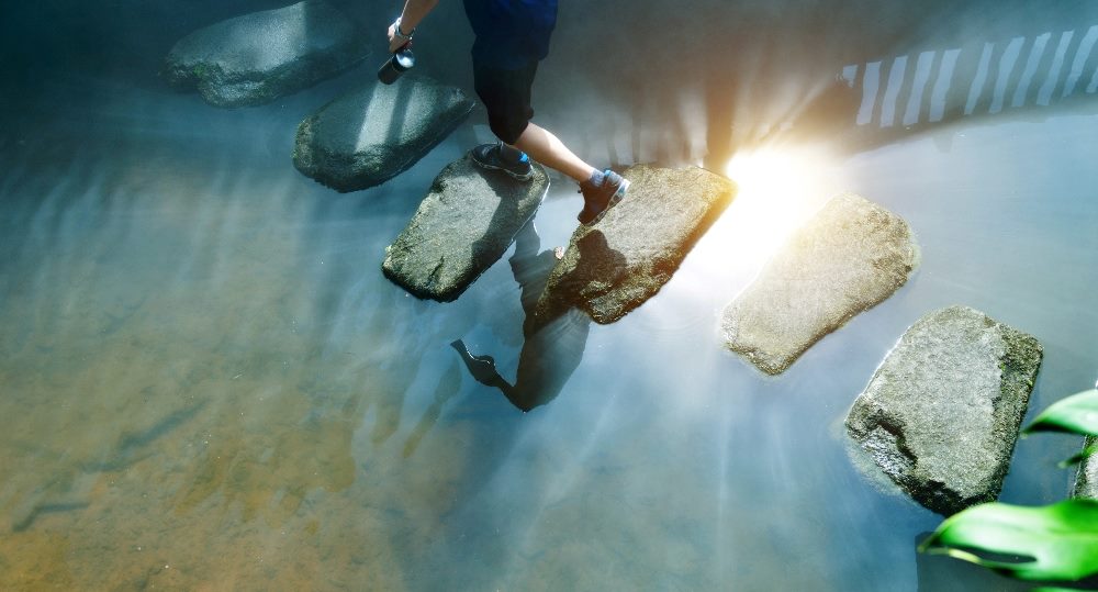 A person wearing shorts and tennis shoes walks across stepping stones to cross a stream in piece about AI in analytics and workflow.