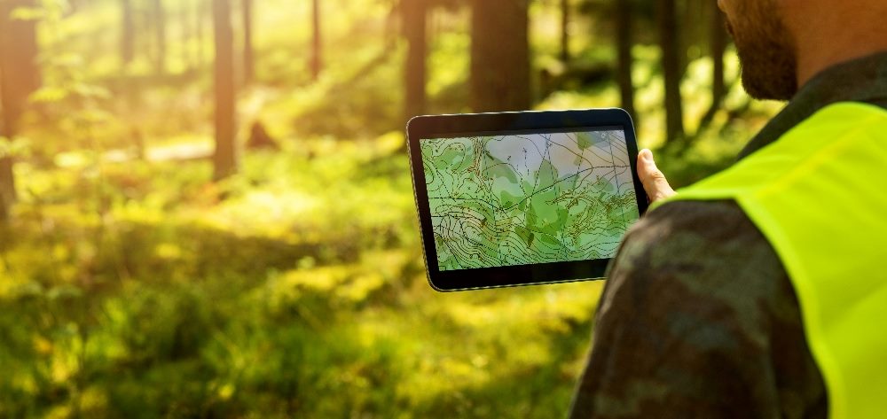 A man holds a tablet with a topographical map visible while holding against the forest landscape in the background, indicating the shifting landscape of digital customer experience software.