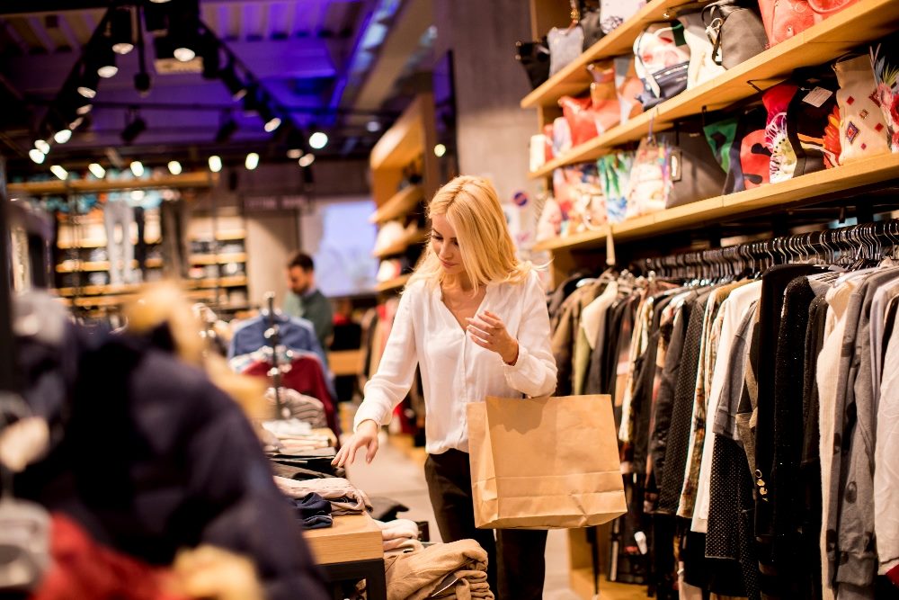A woman with blonde hair, wearing a white blouse, browses through a clothing store. She is holding a paper shopping bag and seems to be looking at various items on sale, with other shoppers visible in the background. The store appears well-lit and modern, featuring a variety of clothing and accessories displayed on racks and shelves in piece about AI customer service.