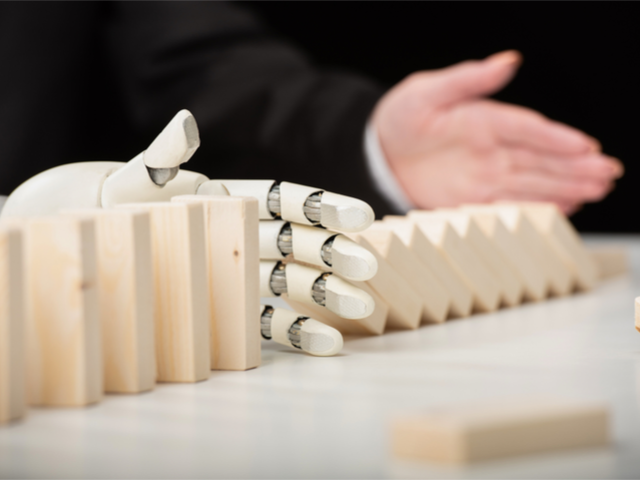 A partial view of a businesswoman pushing wooden dominoes while an AI-driven robotic hand prevents the cascading dominoes - AI challenges concept