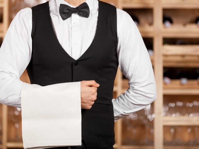 A male waiter in a white shirt, black bow tie and black vest and pants stands ready to serve with a white towel draped over arm on the left and with wooden wine storage racks in the background
