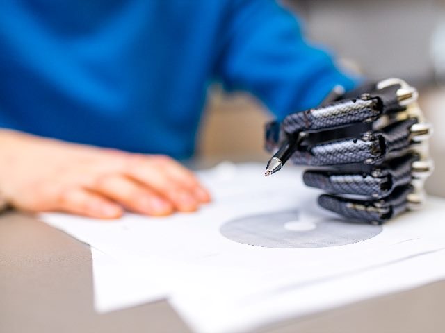 A robot hand with a pen in it begins to write on paper while a human hand holds the paper in place.