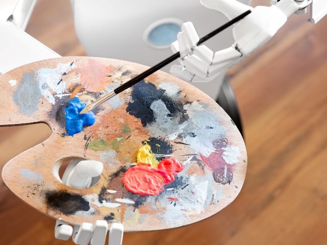 A white robot holds a paint palette with dollops of blue, white, red, yellow, black and gray paint. The robot is dipping his paint brush into the blue paint. 