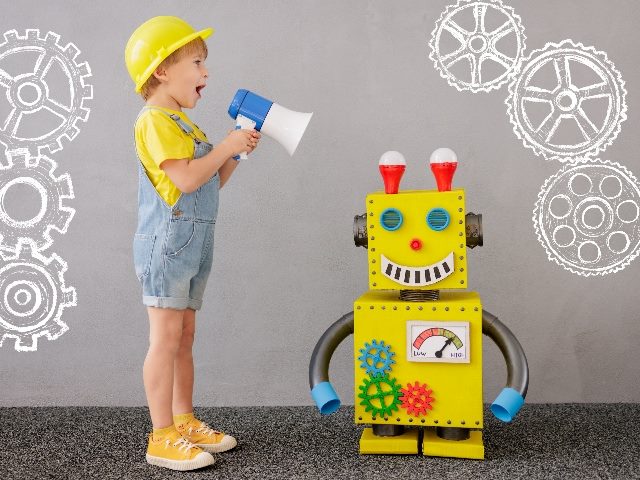 Child standing over a robot wearing a hard hat and speaking into a megaphone. 