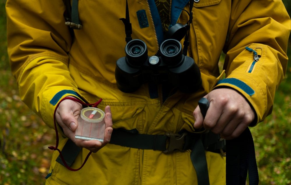 A man wearing yellow jumpsuit is getting ready to go hiking in a forest and holds a compass, binoculars and a walking stick in piece about AI implementation.