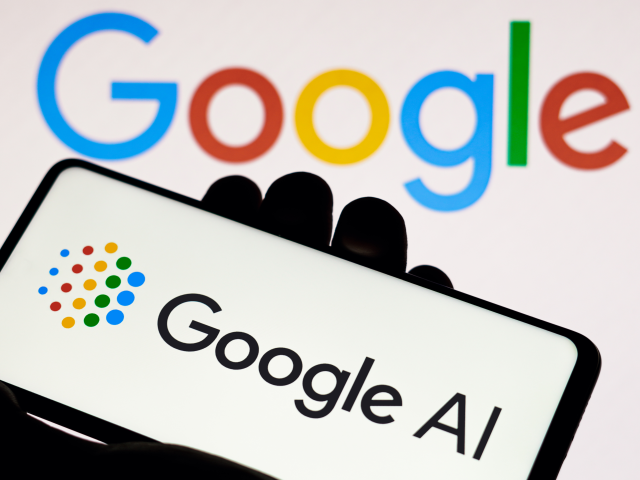 The Google AI logo is displayed on a smartphone screen, in the white background there is an out of focus multi-colored Google logo on the wall