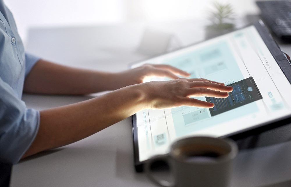 A person is interacting with a large touchscreen device on a desk. The screen displays various data and interface elements, indicating a high-tech environment. Their hands are actively navigating through the content on the screen. There is a coffee mug and a small plant on the desk, creating a modern and minimalistic workspace. The overall scene suggests the use of advanced technology such as AI in customer experience needs the human touch.