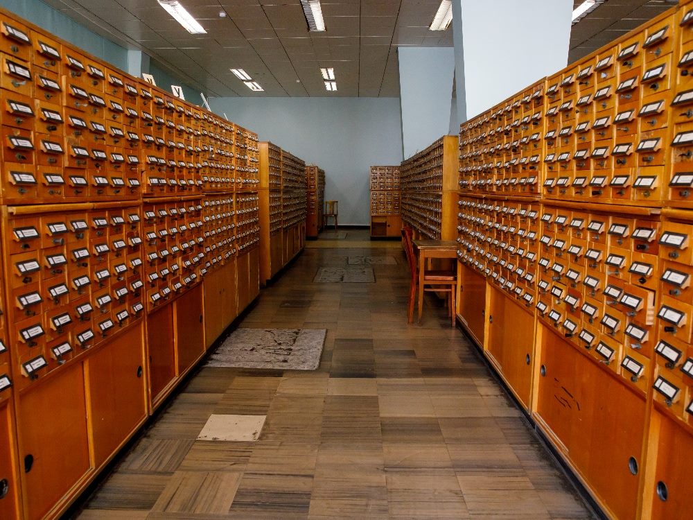 An aisle of ld vintage wooden library card catalog cabinets separated by a tiled floor in piece about AI-powered search.