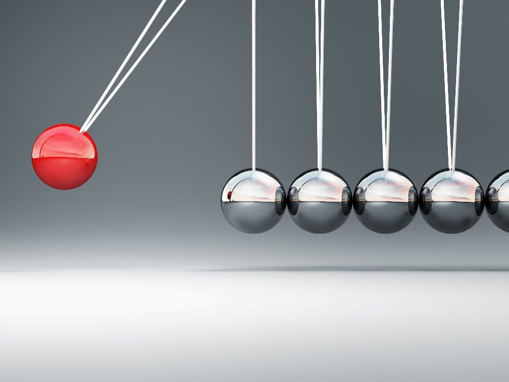 A Newton's Cradle with one red metal ball hanging from a string swinging from the left into five silver metal balls hanging from strings on the right, suggesting the impact of AI on marketing in the next decade.