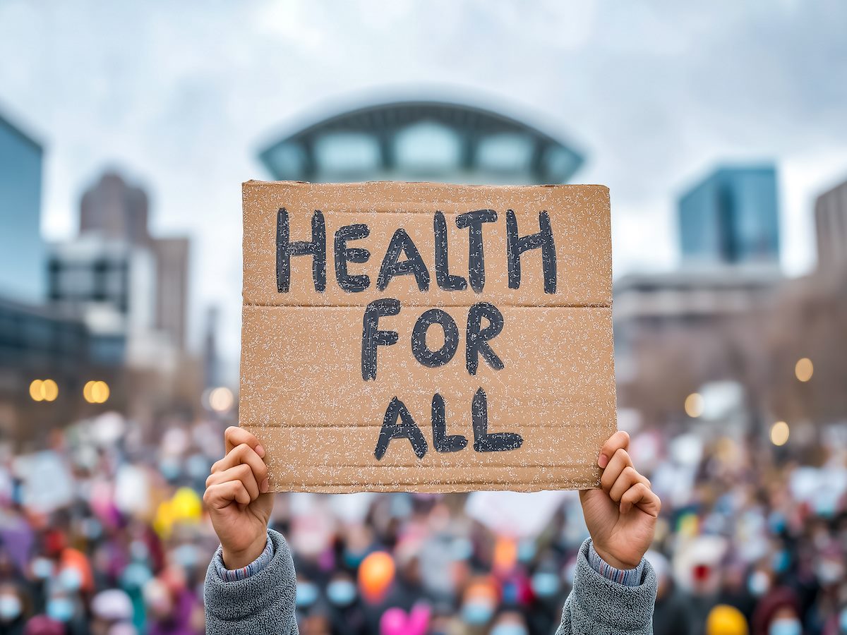 A gathering of people outside, with one person holding a cardboard sign that says "health for all" 