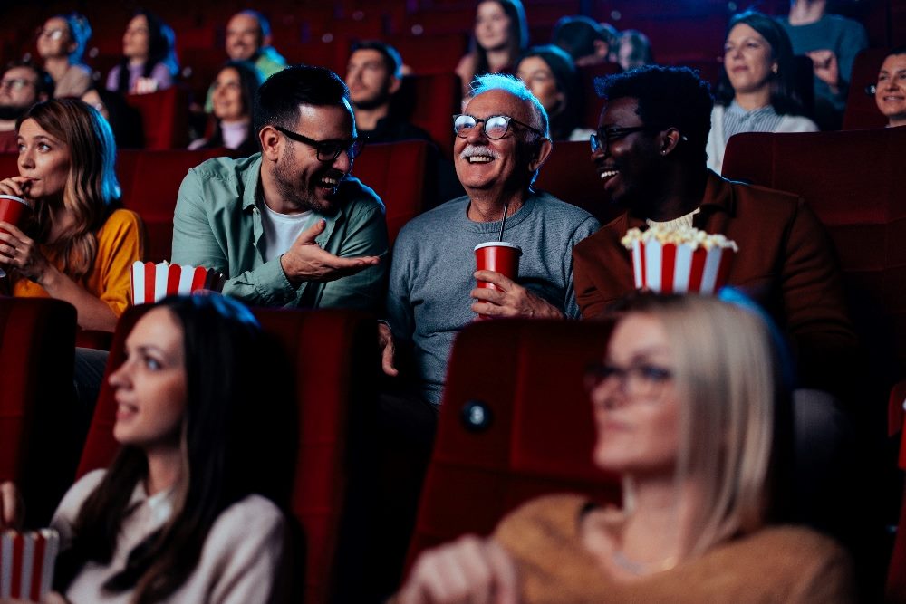 This image shows a diverse group of people enjoying a movie together in a theater. The group includes a man with glasses laughing joyously while holding a cup, seated between two other men—one discussing something with him and another smiling towards him. The audience is engaged and happy, some holding popcorn, and everyone appears to be having a good time. The scene captures a moment of shared entertainment in a cozy, red-seated movie theater in piece about the impact of AI ecommerce, including brand storytelling.