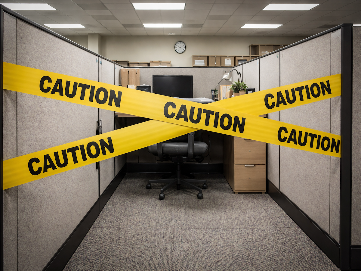 Yellow caution tape crisscrosses the entrance of an empty office cubicle, blocking access to a desk, computer and chair under fluorescent ceiling lights